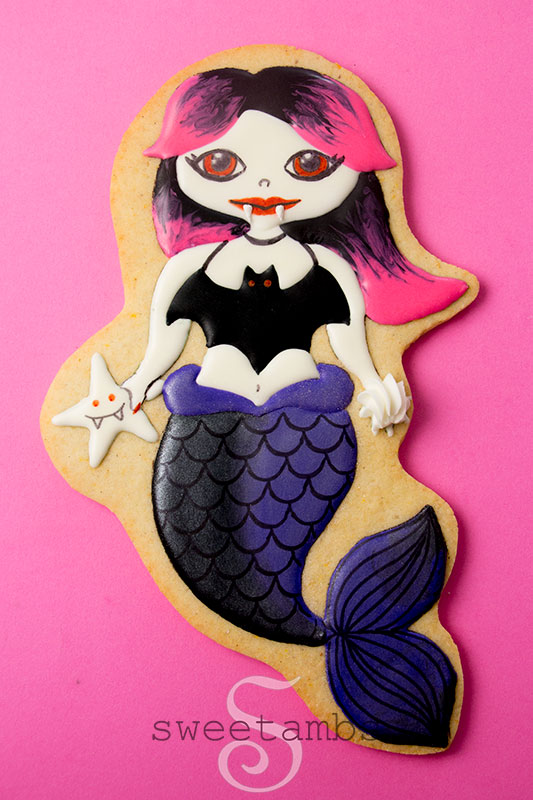 Een koekje dat is versierd om eruit te zien als een vampierzeemeermin met een zwarte en paarse staart en roze en zwart haar.  Het koekje is op een hete roze achtergrond.  De vampiermeermin houdt een vampierzeester in de ene hand en een zeeschelp in de andere.