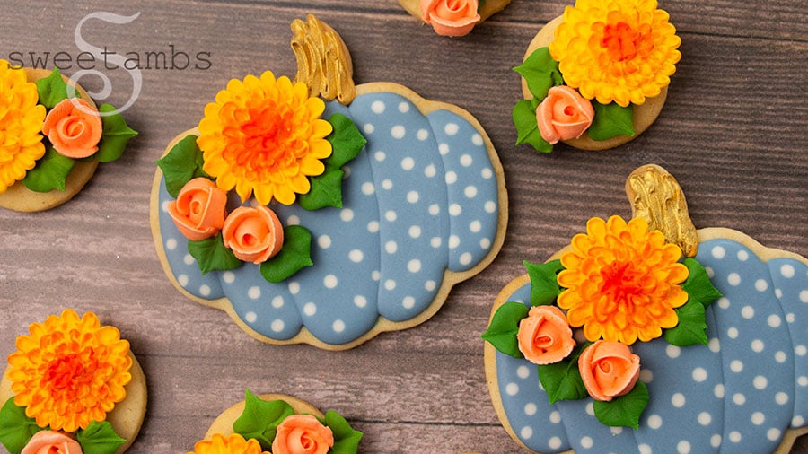 pumpkin shaped cookies decorated with light blue royal icing white white polka dots. The pumpkin cookies have gold stems and are also decorated with royal icing roses and chrysanthemums. The cookies are surrounded by more royal icing flowers and are on a wooden background.