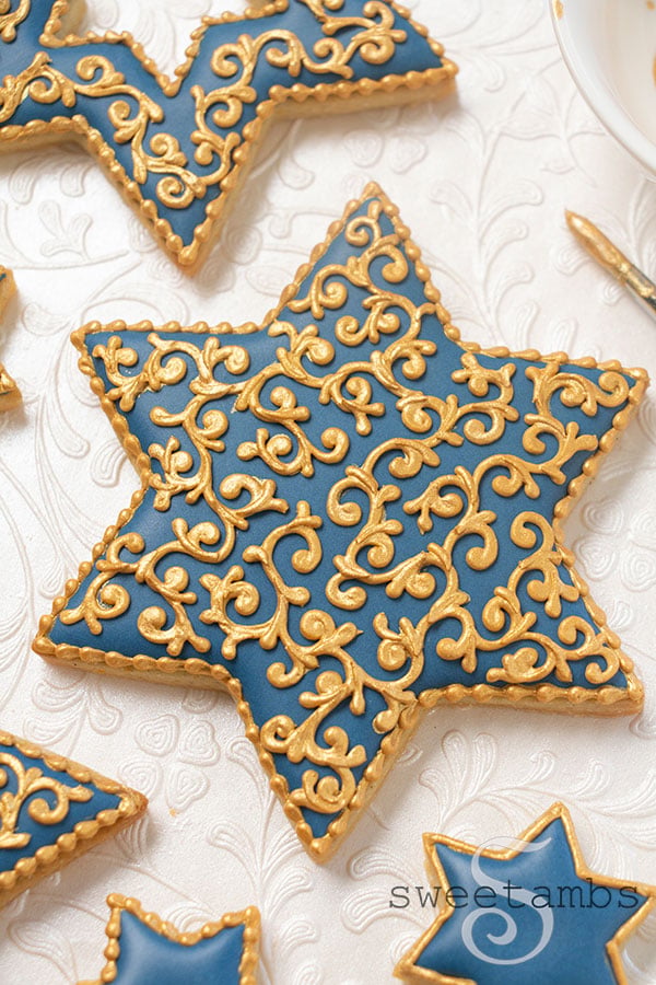 Star of David shaped cookies decorated with blue icing and gold filigree details. The cookies are next to a dish of gold luster dust and a paint brush. The background is pearl white embossed with filigree.