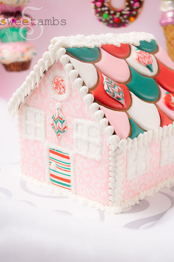 A pink, red, white, and green gingerbread house with a scalloped roof decorated with royal icing ornaments. The house is sitting on a white fluffy surface in front of a pink background. There are ornaments that look like cupcakes, donuts, and ice cream hanging in the background