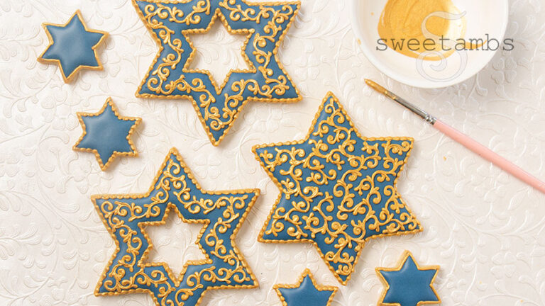 Star of David shaped cookies decorated with blue icing and gold filigree details. The cookies are next to a dish of gold luster dust and a paint brush. The background is pearl white embossed with filigree.