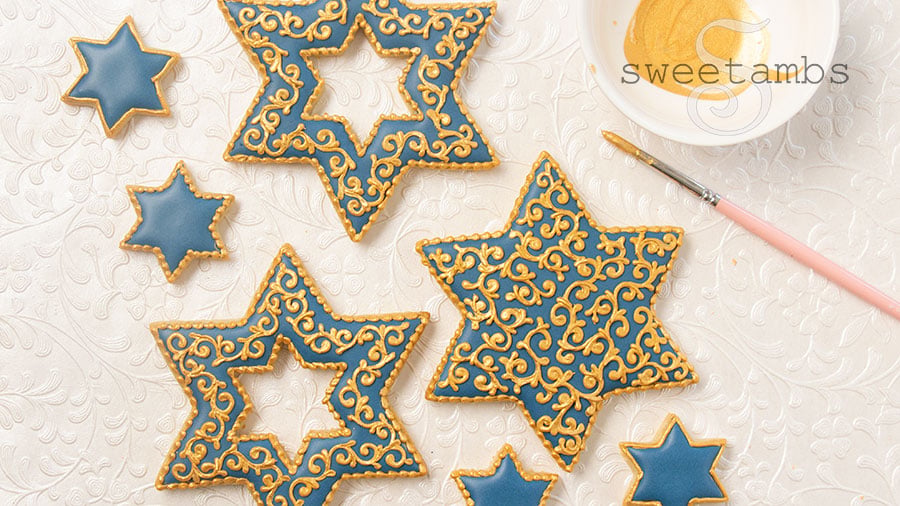 Star of David shaped cookies decorated with blue icing and gold filigree details. The cookies are next to a dish of gold luster dust and a paint brush. The background is pearl white embossed with filigree.