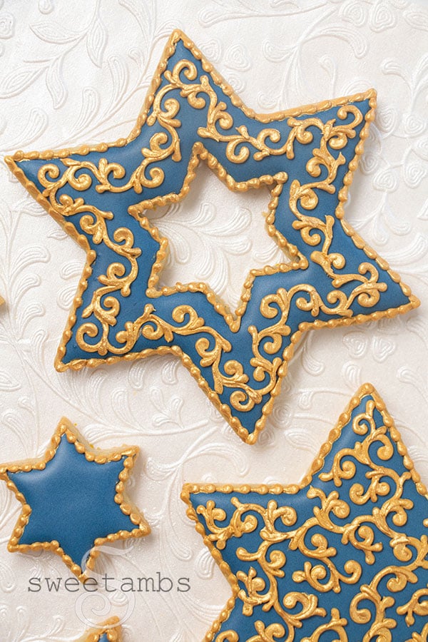 Star of David shaped cookies decorated with blue icing and gold filigree details. The cookies are next to a dish of gold luster dust and a paint brush. The background is pearl white embossed with filigree.