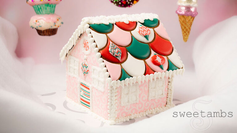 A pink, red, white, and green gingerbread house with a scalloped roof decorated with royal icing ornaments. The house is sitting on a white fluffy surface in front of a pink background. There are ornaments that look like cupcakes, donuts, and ice cream hanging in the background
