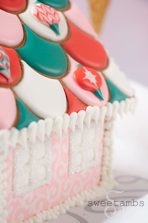 A close up of the scalloped roof and one side of the gingerbread house. The roof is decorated with royal icing ornaments are there are royal icing icicles hanging on the edge of the roof. The windows are covered in white sparkling sugar.