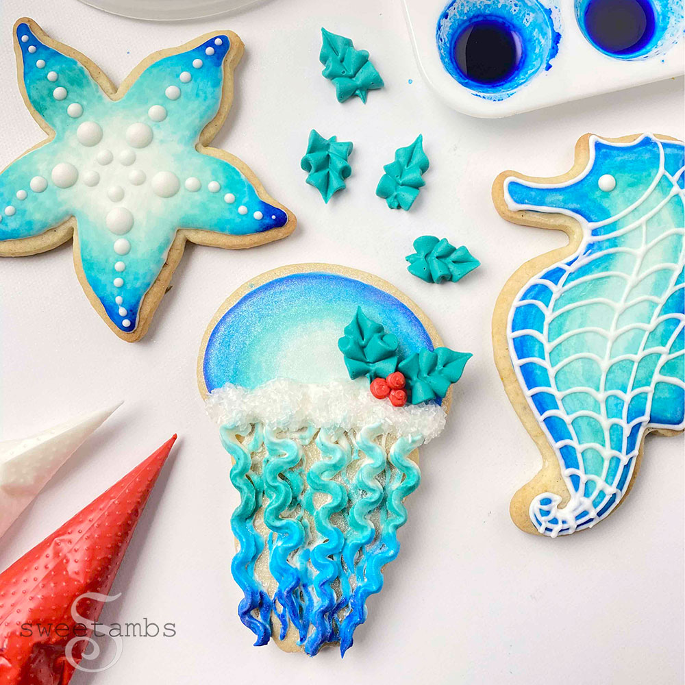 a set of cookies decorated with an under the sea theme with a seahorse, starfish, and jellyfish. The cookies are shades of blue with white sparkling sugar and royal icing holly leaves and berries. There are two piping bags filled with icing and a paint palette with blue food coloring around the cookies.