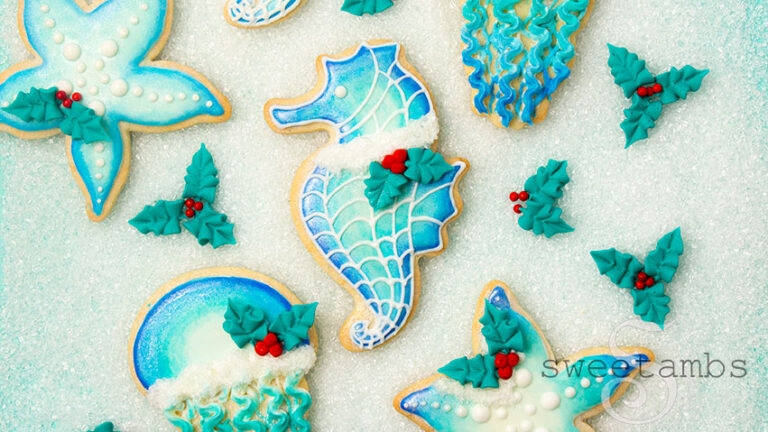 a set of cookies decorated with an under the sea theme with a seahorse, starfish, and jellyfish. The cookies are shades of blue with white sparkling sugar and royal icing holly leaves and berries. The cookies on laying on a layer of sparkling sugar.