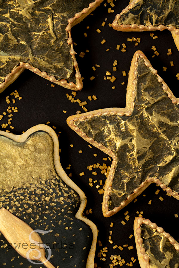 A set of cookies decorated in black and gold royal icing for New Year's Eve. There are black star cookies with gold accents. The cookies are on a black background sprinkled with gold sparkling sugar.