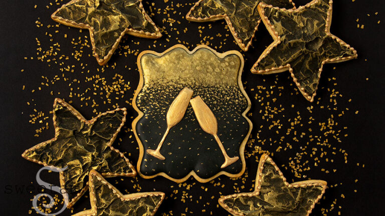 A set of cookies decorated in black and gold royal icing for New Year's Eve. There is a plaque shaped cookie in the middle with two gold Champagne glasses on a gold ombre background on black icing. The cookie is surrounded by black star cookies with gold accents. The cookies are on a black background sprinkled with gold sparkling sugar.