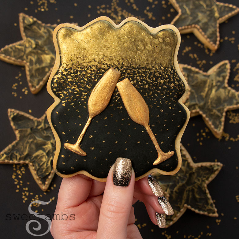 A set of cookies decorated in black and gold royal icing for New Year's Eve. There is a plaque shaped cookie in the middle with two gold Champagne glasses on a gold ombre background on black icing being held by gold ombre and black painted fingernails. The cookie is surrounded by black star cookies with gold accents. The cookies are on a black background sprinkled with gold sparkling sugar.