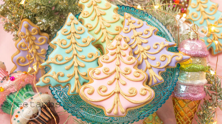 A plate of pastel Christmas tree cookies decorated with a gold filigree design. The cookies are on a teal glass cake platter. There are Christmas decorations in the background including sparkly greenery, a cupcake ornament, and an ice cream cone ornament.