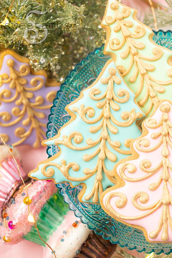 A plate of pastel Christmas tree cookies decorated with a gold filigree design. The cookies are on a teal glass cake platter. There are Christmas decorations in the background including sparkly greenery, and a cupcake ornament.