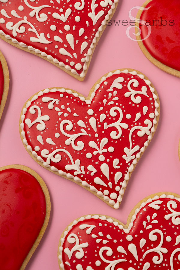 Valentines day cookies decorated with red royal icing and an off-white filigree design. The cookies are on a pink background.