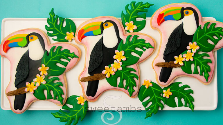 toucan cookies decorated with royal icing are on a white rectangular platter on a teal background. The cookies are surrounded with monstera leaves royal icing transfers.