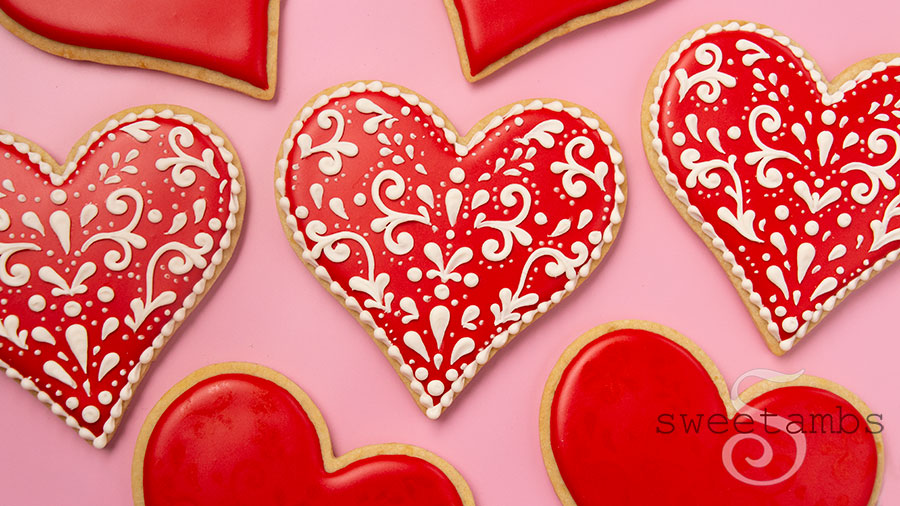 Valentines day cookies decorated with red royal icing and a light beige filigree design. The cookies are on a pink background.