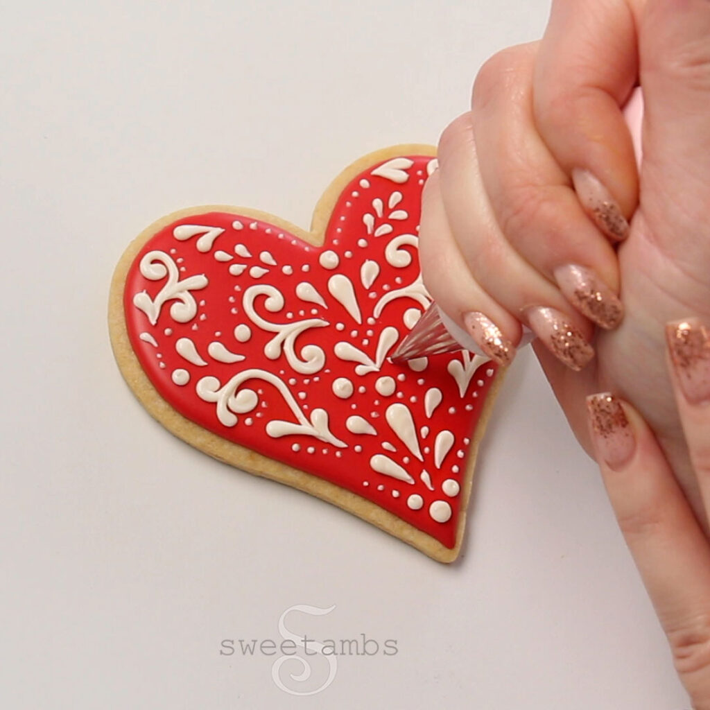 Piping filigree with light beige royal icing on a red heart shaped cookie