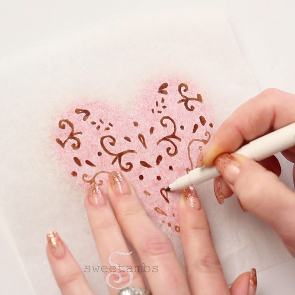 Tracing a template with an edible ink marker on a piece of tissue paper over an iced heart shaped cookie.