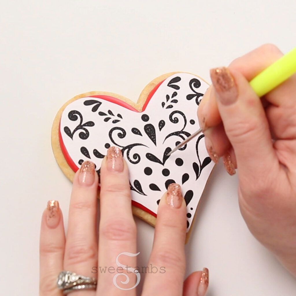 Poking holes in a template with a scribe tool to transfer a template onto an iced heart shaped cookie