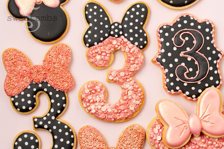 A set of cookies decorated with rose gold and black and white polka dot royal icing. The cookies are shaped like the number 3 with bows on top. There is also a plaque cookie with the number 3 in royal icing.