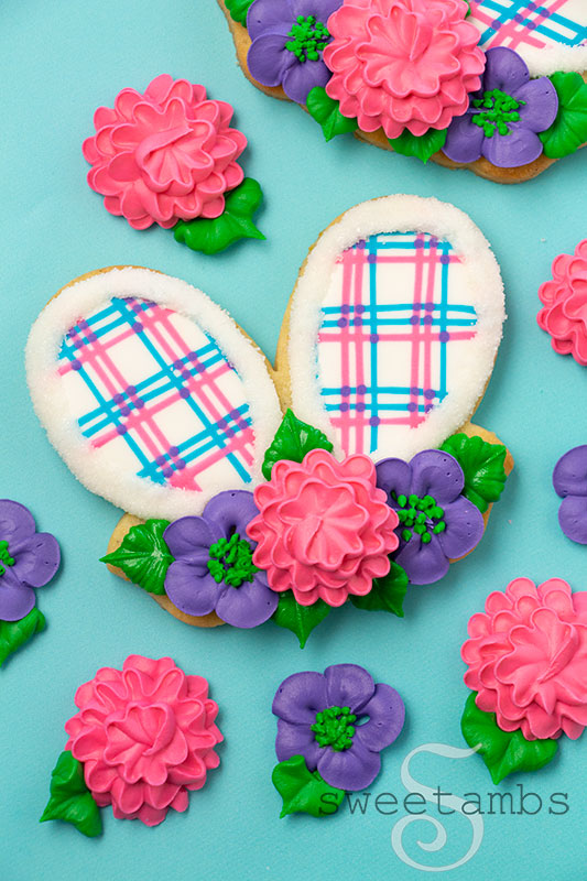 easter bunny ears cookies decorated with brightly colored plaid royal icing and royal icing flowers. The cookies are on a light blue background.