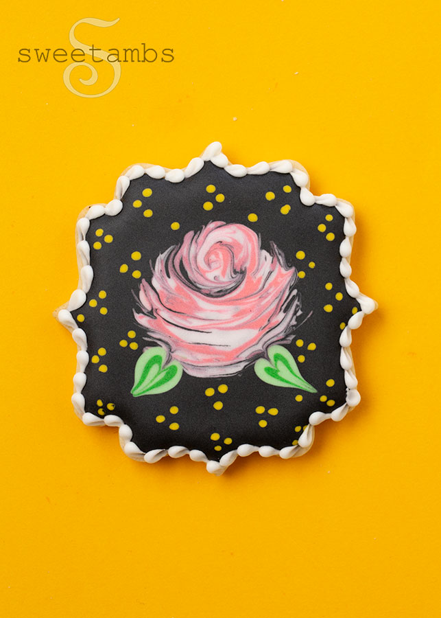 A plaque cookie decorated with one large rose surrounded by yellow dots on black royal icing. The cookie is on a golden yellow background.