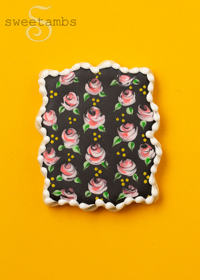 A plaque cookie decorated with small pink roses and yellow dots on black royal icing. The cookie is on a golden yellow background.