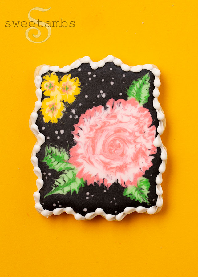 A plaque cookie decorated with pink and yellow flowers on black royal icing. The cookie is on a golden yellow background.