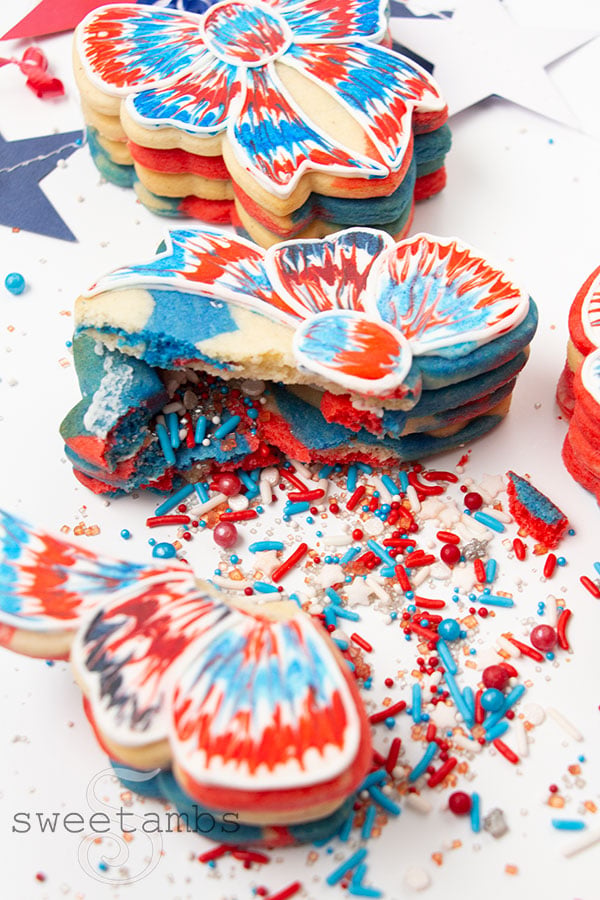 A bow shaped cookie decorated with red, white, and blue royal icing in a tie dye effect broken open with sprinkles spilling out