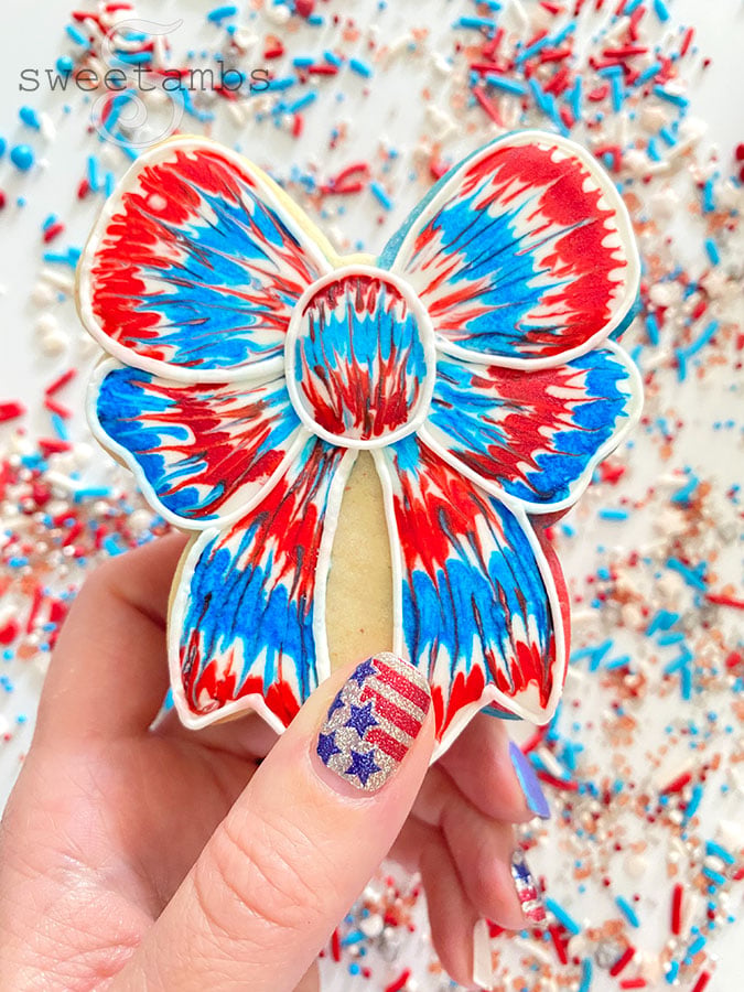 A bow shaped cookie decorated with red, white, and blue royal icing with a tie dye effect being held by a hand with red, white, and silver nail polish with stars and stripes. There are red, white, and blue sprinkles in the background.