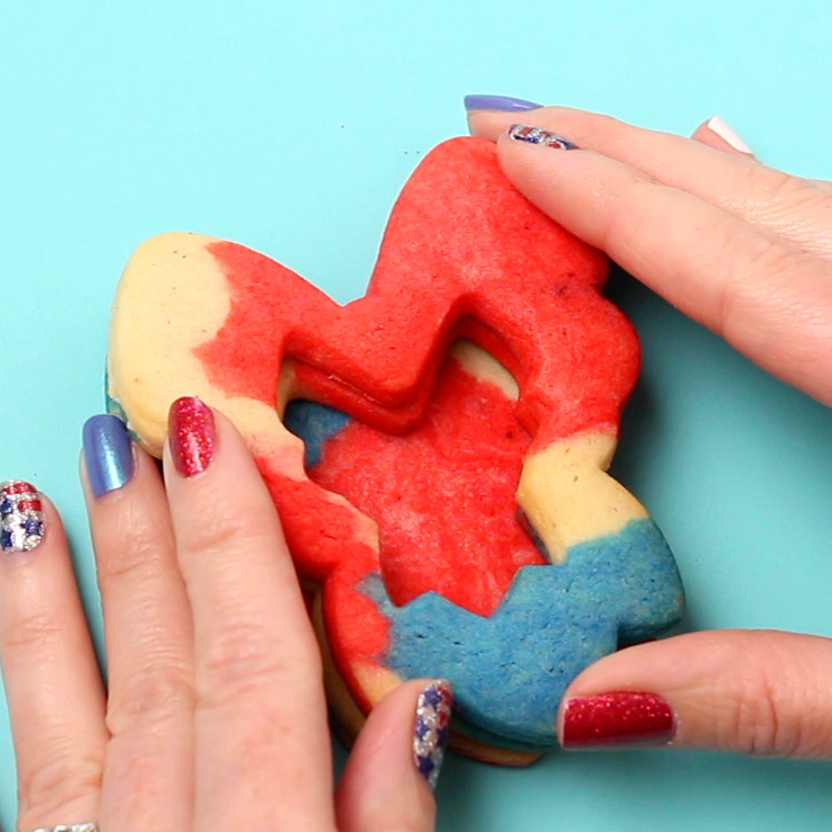 Hands assembling red, white, and blue bow shaped cookies with a well in the center. The fingernails are decorated with red, blue, and silver nail polish with Stars and Stripes.