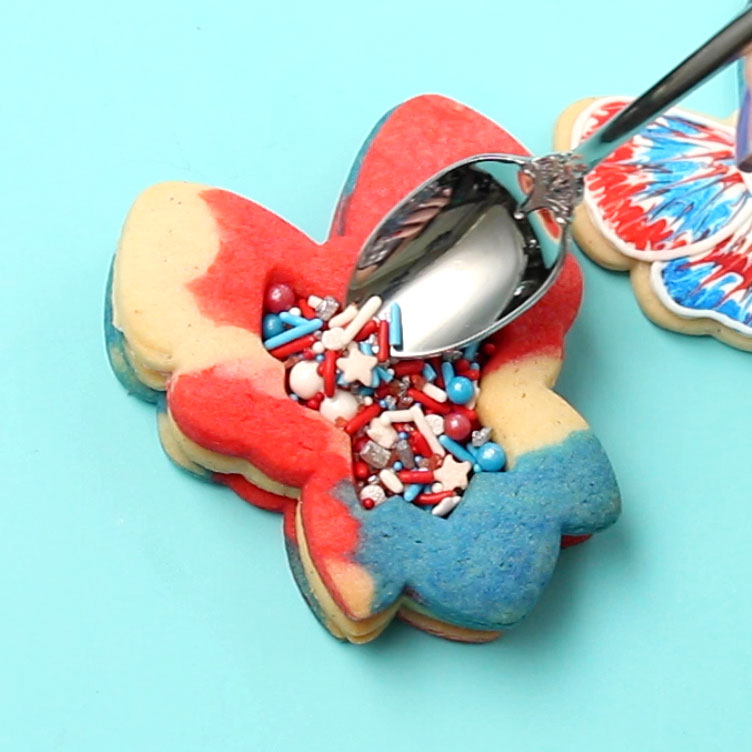 A spoon filling a sugar cookie with red, white, and blue sprinkles. The cookie is shaped like a bow and is marbled with red, white, and blue.