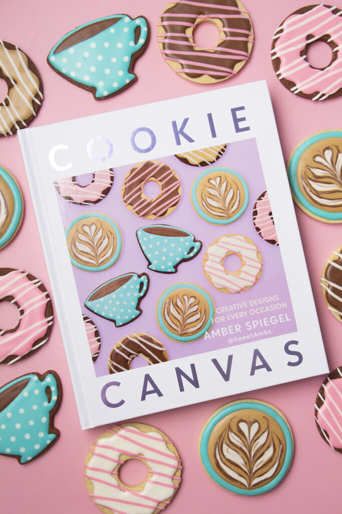 A hardcover book surrounded by cookies decorated to look like coffee, lattes, and donuts. The cover of the book show an image of the same cookies. The title of the book is Cookie Canvas. The subtitle reads creative designs for every occasion Amber Spiegel @sweetambs