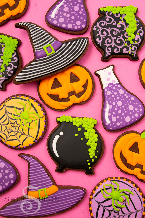 Brightly colored Halloween cookies decorated to look like a jack-o'lantern wearing a striped witch hat, spider webs with neon green spiders, a bubbling cauldron, and a science beaker