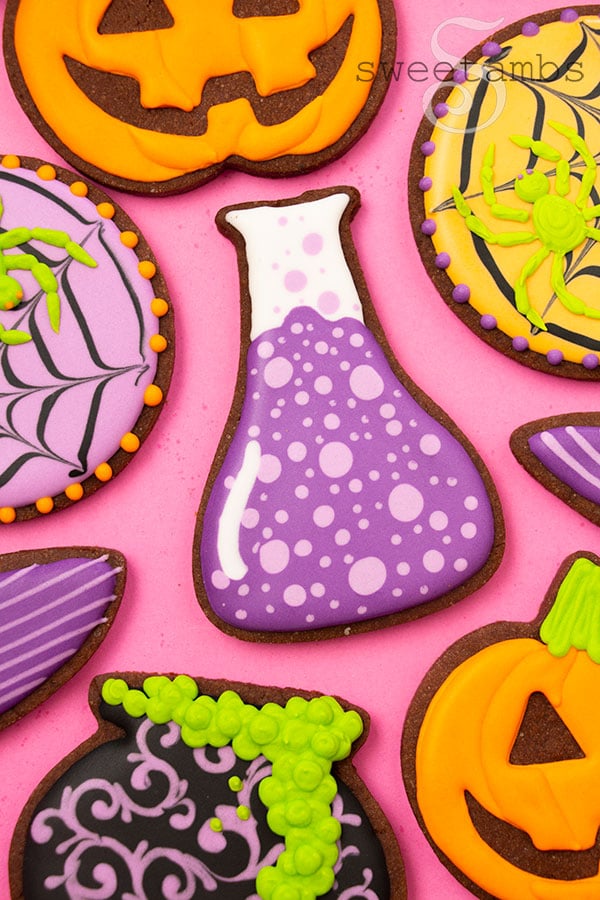 A science beaker cookie decorated to look like it's filled with a bubbling purple liquid. The cookie is surrounded by brightly colored Halloween cookies