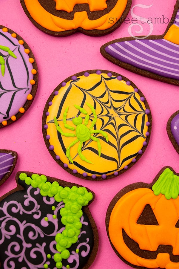 An orange iced cookie decorated to look like a spider web with a neon green spider on it. The cookie is surrounded by brightly colored Halloween cookies.