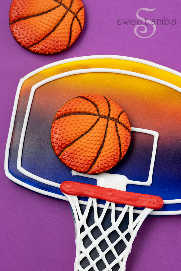 A basketball cookie on a backboard cookie. The cookies are decorated with royal icing. The backboard cookie has a yellow, orange, pink, and blue design resembling a sunset. The basketball cookies are shaded around the edges to give a 3 dimensional look.
