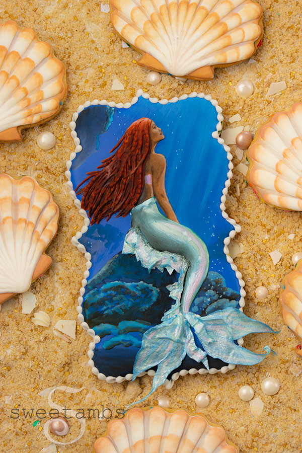 A plaque shaped cookie decorated to look like Ariel sitting on a rock in the ocean from the live action version of The Little Mermaid