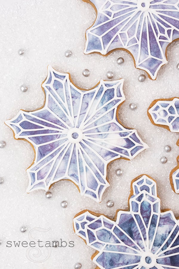 Snowflake shaped cookies decorated with a blue and purple watercolor design with white snowflake outlines on top. The cookies are on a white sugar background with silver sugar pearls in between the cookies.