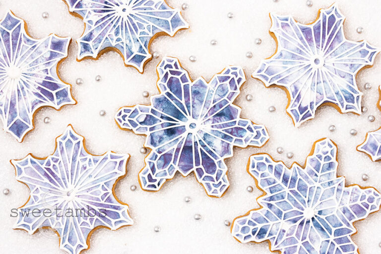 Snowflake shaped cookies decorated with a blue and purple watercolor design with white snowflake outlines on top. The cookies are on a white sugar background with silver sugar pearls in between the cookies.