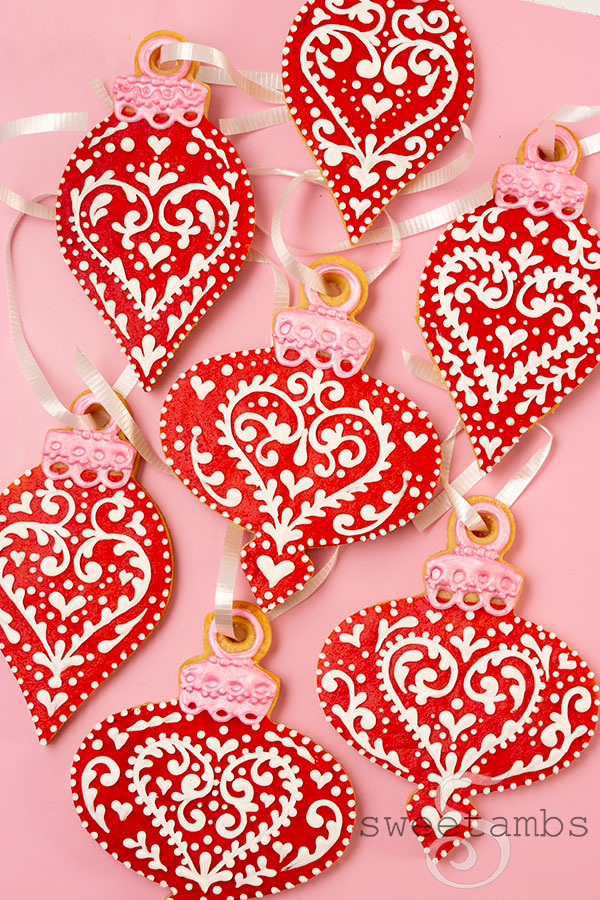 A set of red ornament shaped cookies decorated with a white filigree design. The ornament cookies have white curling ribbon strung through the top.