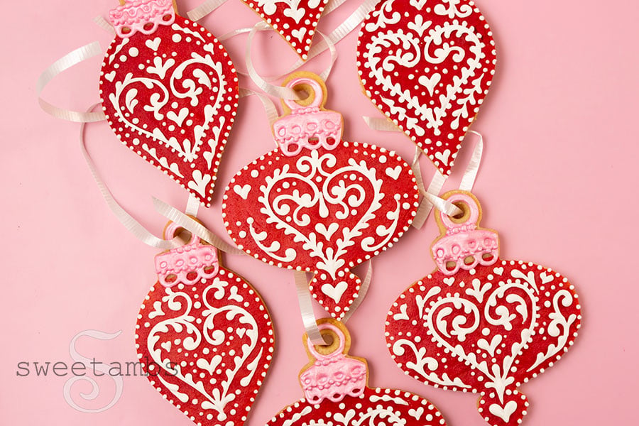 A set of red ornament shaped cookies decorated with a white filigree design. The ornament cookies have white curling ribbon strung through the top.
