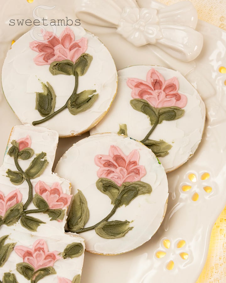 A set of round cookies decorated with flowers created with a palette knife painting technique. The cookies are on a platter with an eyelet lace pattern and a ceramic bow over a lace tablecloth with glowing fairy lights underneath