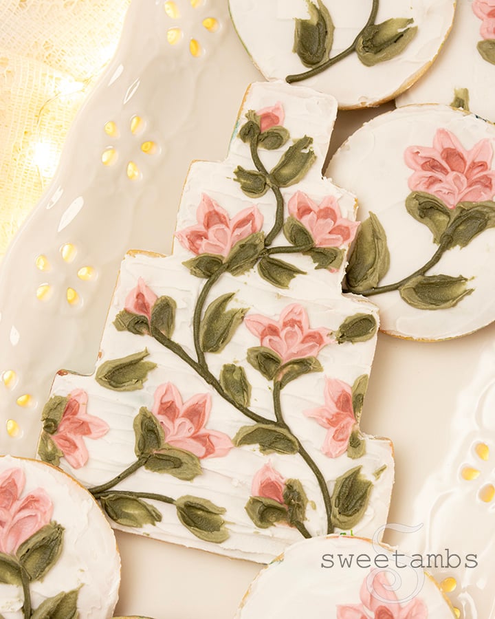 A cookie decorated to look like a wedding cake with flowers created with a palette knife painting technique. The wedding cake cookie is surrounded by smaller round cookies with flowers. The cookies are on a platter with an eyelet lace pattern over a lace tablecloth with glowing fairy lights underneath