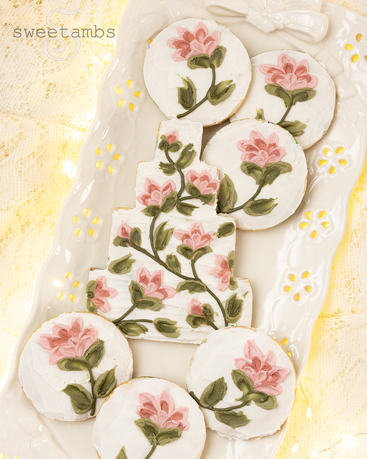 A cookie decorated to look like a wedding cake with flowers created with a palette knife painting technique. The wedding cake cookie is surrounded by smaller round cookies with flowers. The cookies are on a platter with an eyelet lace pattern over a lace tablecloth with glowing fairy lights underneath