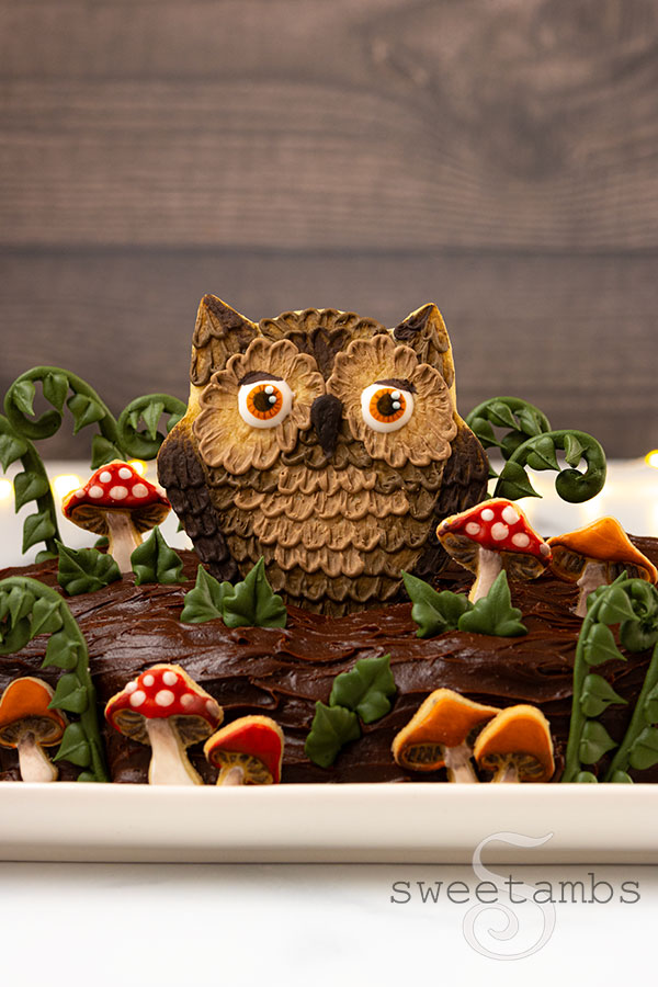 A cake decorated to look like a log with fairytale mushrooms, ferns, leaves, and an owl cookie