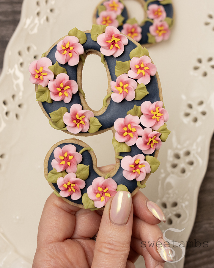 A hand with shimmery light pink nails holding a number 9 cookie decorated with dark blue icing and royal icing cherry blossoms. There is a platter in the background with an eyelet lace design along the edges.