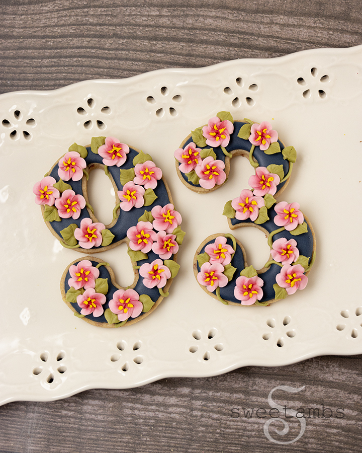 Cookies shaped like the number 93 decorated with dark blue icing and royal icing cherry blossoms. The cookies are on a platter with an eyelet lace design over a wooden background.