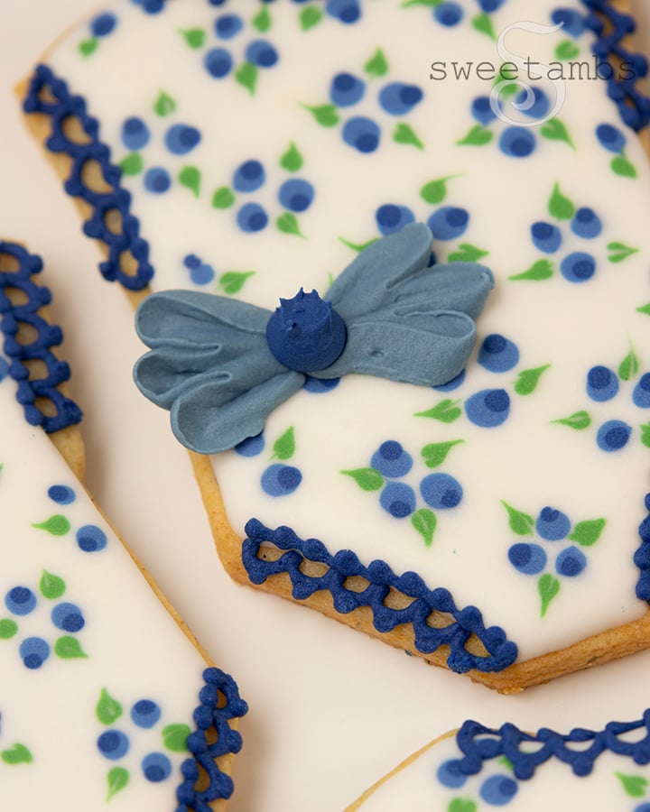 A close up of a cookie decorated to look like a baby romper with a blueberry pattern and a royal icing bow with a royal icing blueberry in the center.