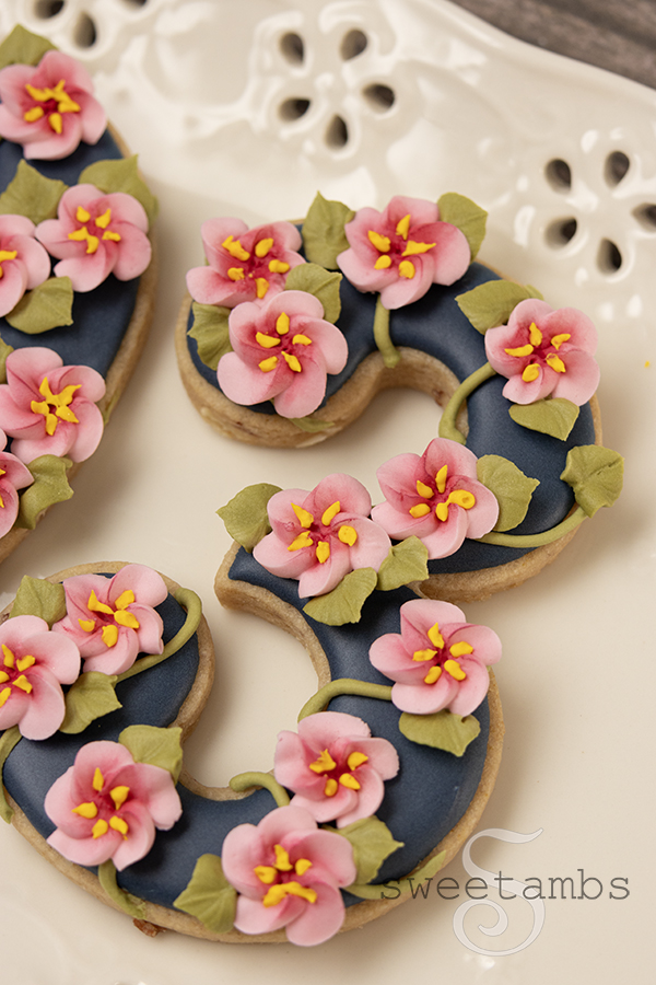 A cookie shaped like the number 3 decorated with dark blue icing and royal icing cherry blossoms. The cookie is on a platter with an eyelet lace design.