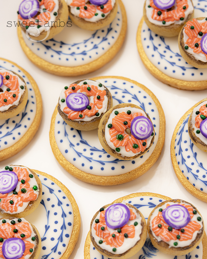 Cookies decorated to look like tiny bagels topped with cream cheese, lox, onions, and capers. The bagel cookies are on decorated cookies decorated to look like a plate with a blue and white pattern.